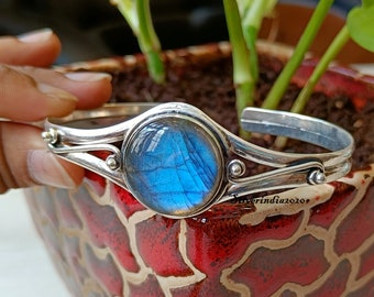 Puede incluir: Un brazalete de plata con una gran piedra redonda de labradorita azul engastada en el centro. El brazalete tiene un diseño simple y elegante con dos bandas que se envuelven alrededor de la muñeca.
