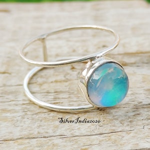 Peut inclure: Une bague en argent ornée d'une grosse pierre d'opale ronde et irisée. La bague présente un design unique à double bande. L'opale affiche des nuances de bleu, de vert et de blanc. La bague est posée sur une surface en bois.