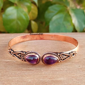Op de afbeelding: Koperkleurige open manchetarmband met twee druppelvormige, dieppaarse edelstenen. De armband heeft sierlijk, gedetailleerd metaalwerk rond de edelstenen en langs de zijkanten. De armband wordt gepresenteerd op een houten oppervlak.