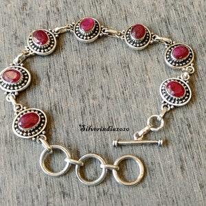 Puede incluir: Una pulsera de plata con cierre de palanca presenta siete piedras preciosas rojas de forma ovalada engastadas en engastes de plata. Cada engaste está rodeado por una fila de pequeñas cuentas de plata.