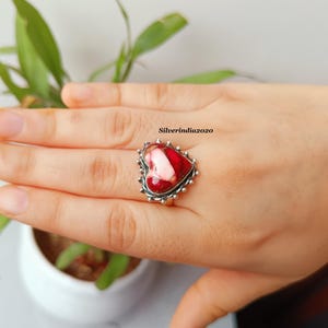 May include: A heart-shaped ring with a red and white stone set in a silver band. The ring is on a hand, with a plant in the background. The ring has a textured border and is a piece of jewelry.