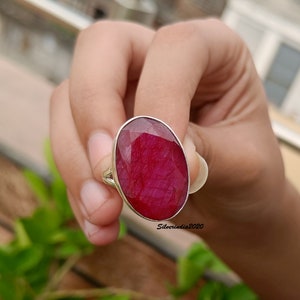 Pode incluir: Um anel de prata com uma grande pedra preciosa de rubi de corte oval. O rubi &eacute; de cor vermelha profunda e est&aacute; engastado numa banda de prata. O anel &eacute; segurado contra um fundo de folhagem verde.