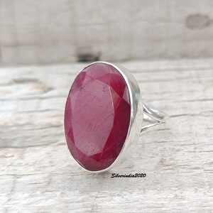 Pode incluir: Um anel de prata com uma grande gema de rubi de corte oval. O rubi &eacute; de cor vermelha profunda. O anel tem um design de haste dividida. O fundo &eacute; uma superf&iacute;cie de madeira de cor clara.