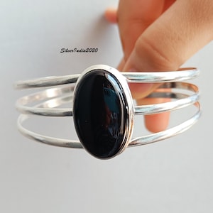 May include: Silver cuff bracelet with a large, oval, black onyx stone. The bracelet has three parallel bands and a polished finish. The jewelry is held by a hand against a light background. The text "SilverIndia2020" is visible.