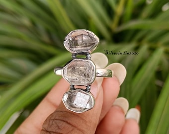May include: A silver ring with three clear, faceted gemstones set in a vertical row. The ring is on a finger.