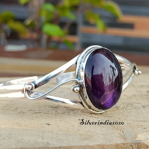 Könnte beinhalten: Ein silbernes Armreif-Armband mit einem großen, ovalen Amethyst-Edelstein. Das Armband hat ein zartes, gedrehtes Design und ist mit kleinen, runden Perlen verziert.