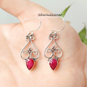 May include: A pair of silver-toned drop earrings with a teardrop-shaped, deep red gemstone. The earrings feature an ornate, swirling design with a hook closure. The text "Silverindia2020" is visible in the background.
