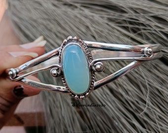 May include: Silver bangle bracelet with a light blue oval gemstone set in a silver bezel. The bracelet has a delicate, intricate design with three bands of silver.