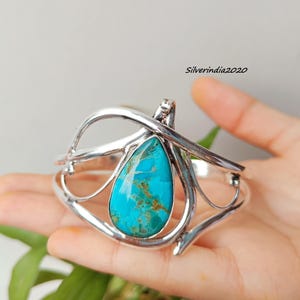 May include: A silver cuff bracelet featuring a large, teardrop-shaped turquoise stone. The stone is a vibrant blue-green color with brown matrix patterns. The bracelet has an open design with curved silver accents.