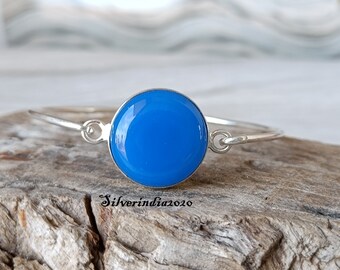 Könnte beinhalten: Ein silbernes Armreif-Armband mit einem leuchtend blauen runden Cabochon-Edelstein. Das Armband ist offen und hat einen Hakenverschluss.