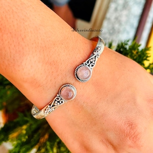 Könnte beinhalten: Silberner Armreif mit zwei rosafarbenen Rosenquarzsteinen. Das Armband hat ein zartes Blumenmuster.