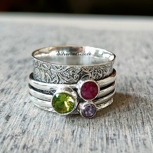 May include: A silver spinning ring with three gemstones. The ring has a textured band with a floral design. The gemstones are green, red, and purple.