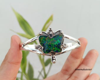 May include: A silver cuff bracelet featuring a central butterfly-shaped inlay with iridescent green and black flecks. The metal band has a split design and small decorative silver spheres at the top and bottom of the butterfly motif.