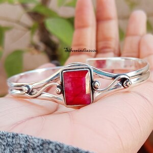 May include: A silver bangle bracelet featuring a red gemstone set in a rectangular frame. The bracelet has a delicate, swirling design.
