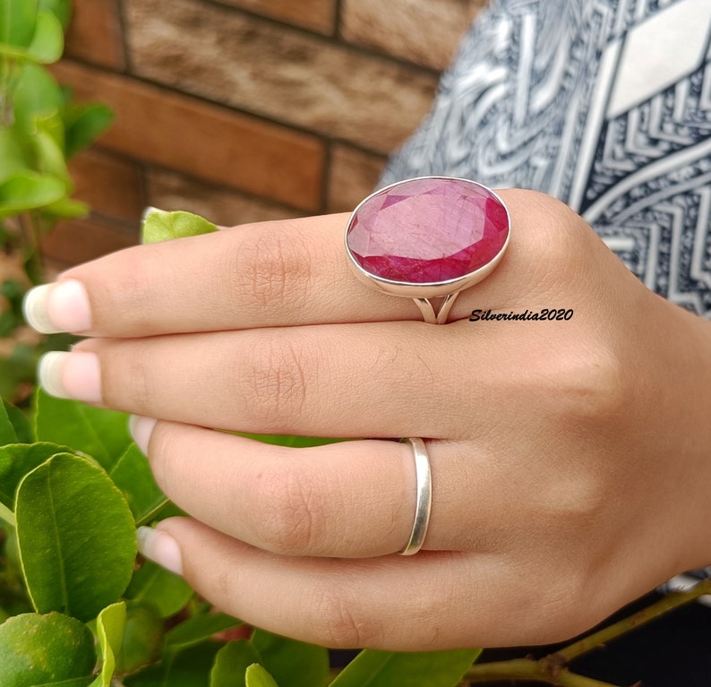 Pode incluir: Um anel de prata com uma grande gema de rubi de corte oval. O rubi &eacute; de cor vermelha profunda e est&aacute; engastado numa moldura de prata simples e elegante. Um anel de banda de prata simples tamb&eacute;m &eacute; vis&iacute;vel. O texto "Silverindia2020" est&aacute; na imagem.