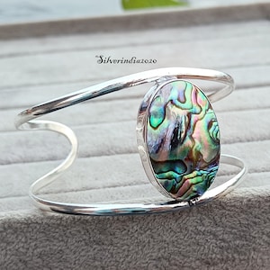 Może przedstawiać: Srebrna bransoletka z dużym owalnym wkładką z muszli abalone. Muszla ma tęczowe zielone, niebieskie i różowe kolory.