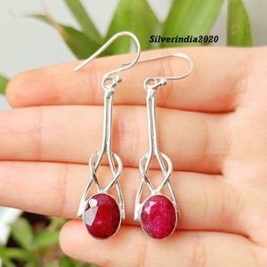 May include: A pair of silver-toned dangle earrings featuring oval-cut, deep red gemstones. The earrings have a unique design with a looped detail above the gemstone and a hook closure. The text "Silverindia2020" is visible.