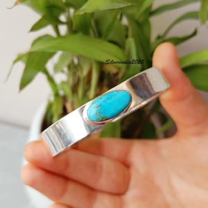 Peut inclure: Un bracelet manchette en argent avec une pierre turquoise vive. Le bracelet rectangulaire a une finition polie et une pierre turquoise ovale centrale. Le bracelet est tenu dans une main, avec un feuillage vert en arrière-plan.