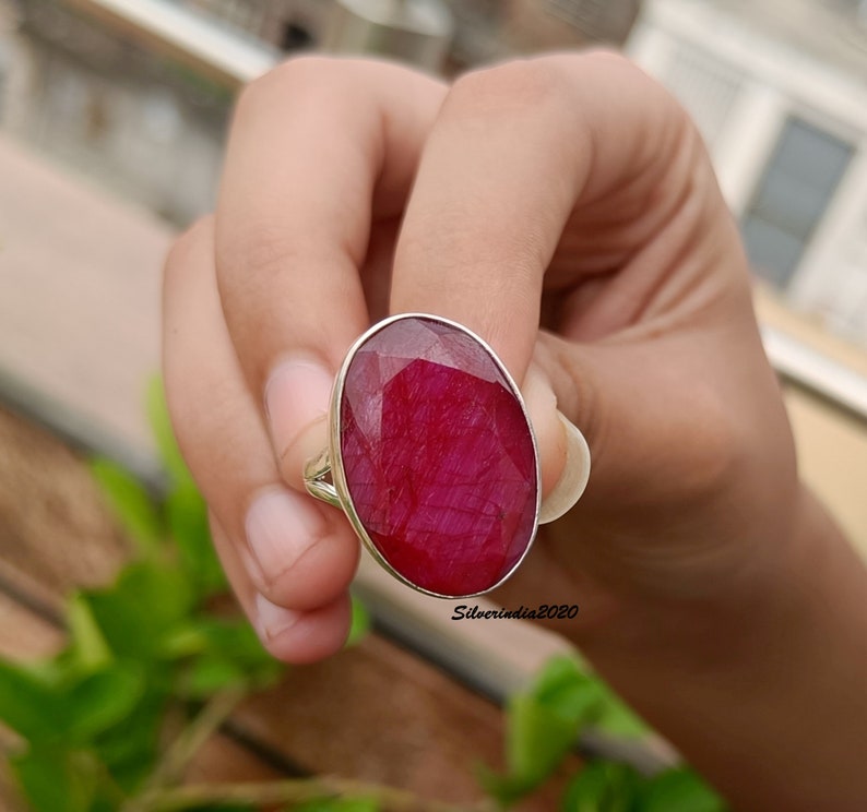 Pode incluir: Um anel de prata com uma grande pedra preciosa de rubi de corte oval. O rubi &eacute; de uma cor vermelha profunda e vibrante e est&aacute; engastado numa simples banda de prata. O anel &eacute; segurado contra um fundo de folhagem verde.