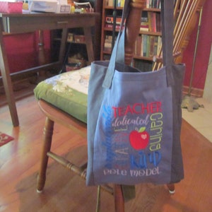 May include: Blue tote bag with an embroidered design that says "Teacher" and "Apple a day keeps the doctor away" with a red apple in the middle. The bag is sitting on a wooden chair.