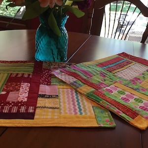 May include: Two colorful patchwork placemats with a yellow border. The placemats are made of different fabrics in shades of red, green, yellow, pink, and white. The placemats are on a wooden table.