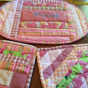 May include: Set of three pink and green quilted placemats with a floral pattern. The center placemat is oval shaped and has the word "Food" embroidered on it.