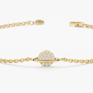 May include: A gold chain bracelet with a small, round, diamond-encrusted charm in the center. The charm is shaped like a planet with a ring around it.