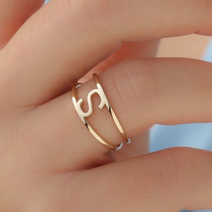 May include: A gold ring with a letter 'S' cut out of the band. The ring is worn on a finger.
