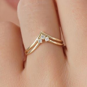 May include: A gold-colored ring with a chevron design. The ring features two parallel bands and a V-shaped top adorned with three small, clear gemstones. The ring is on a finger.
