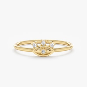 May include: A delicate gold ring featuring an eye design with a central diamond and three smaller diamonds above. The ring has a thin band and a polished finish, set against a white background.