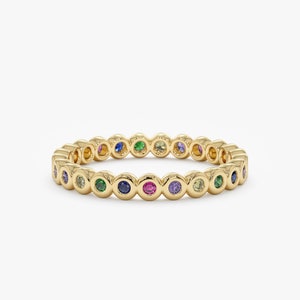 May include: A gold band ring with a row of colorful gemstones. The gemstones are set in a bezel setting and are arranged in a rainbow pattern.