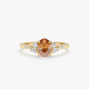 Op de afbeelding: Een gouden ring met een ovale, amberkleurige edelsteen in het midden, omringd door clusters van kleinere, heldere edelstenen. De ring heeft een eenvoudige, gepolijste gouden band.