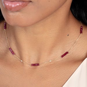 May include: A delicate gold necklace featuring alternating clusters of small, deep red gemstones and tiny, sparkling white stones. The necklace is worn around the neck, showcasing its elegant design and subtle sparkle.