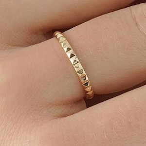 May include: A gold ring with a textured design of small, evenly spaced triangles. The ring is worn on a finger, showcasing its delicate and detailed craftsmanship. The ring's warm gold color contrasts with the skin tone, highlighting its elegant simplicity.