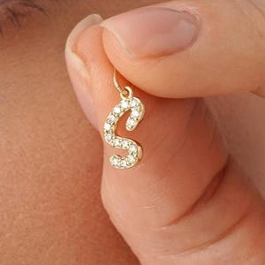 Op de afbeelding: Een goudkleurige bedel in de vorm van de letter "S", bezet met kleine, heldere edelstenen. De bedel wordt door de vingers van een persoon omhoog gehouden, met het oog en een deel van het gezicht zichtbaar op de achtergrond. De bedel is waarschijnlijk voor een ketting of armband.