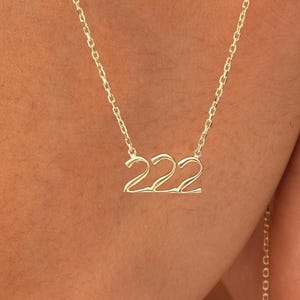 以下が含まれることがあります： 数字の「222」がペンダントになった繊細なゴールドカラーのネックレス。ペンダントは細いチェーンに繋がっています。ニュートラルな背景にネックレスが表示されています。