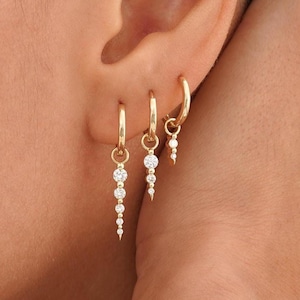 Op de afbeelding: Drie gouden oorringen met bungelende witte edelstenen. De oorbellen hebben verschillende lengtes, waarbij de langste een reeks afgestudeerde stenen heeft. De oorbellen worden op een oor gedragen, wat het ontwerp en de glans van de sieraden laat zien.