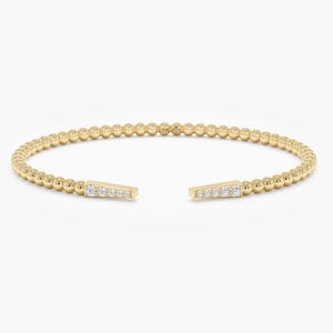May include: A gold-colored open cuff bracelet. The bracelet is made of small, round beads. The ends of the bracelet feature a rectangular design with small, clear stones. The bracelet is set against a white background.