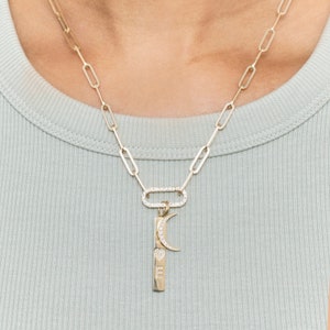 May include: A gold chain necklace with a charm featuring a crescent moon and a small heart with the word "love" inscribed on it. The charm is attached to a larger oval-shaped charm with small diamonds around the edge.