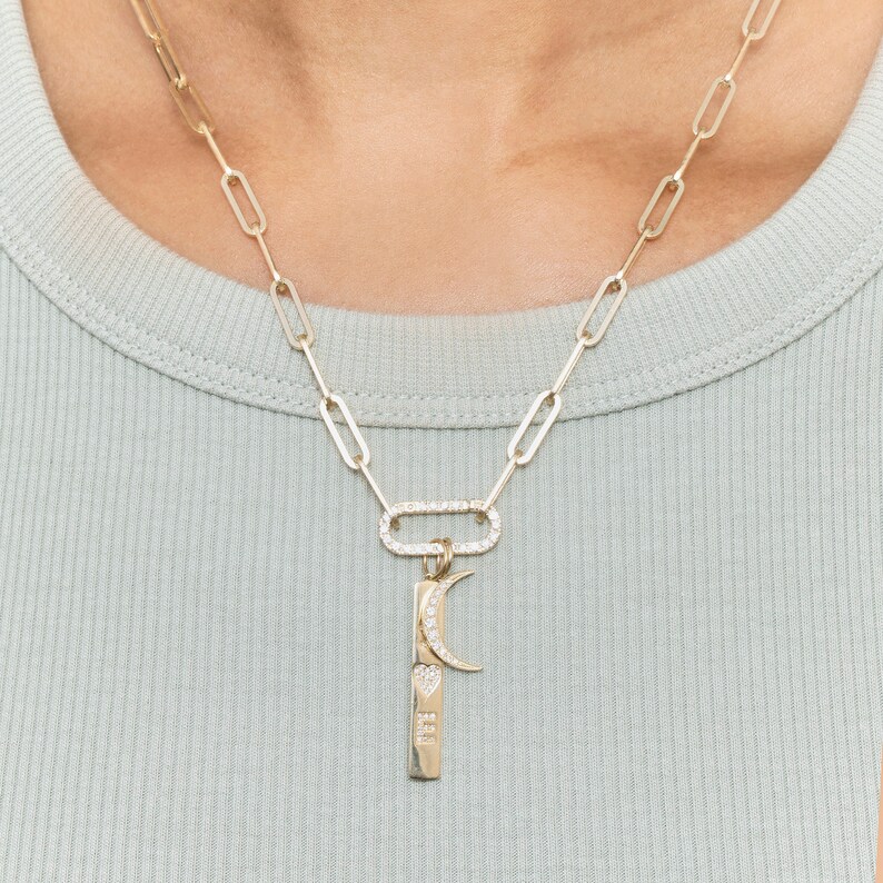 May include: A gold chain necklace with a charm featuring a crescent moon and a small heart with the word "love" inscribed on it. The charm is attached to a larger oval-shaped charm with small diamonds around the edge.