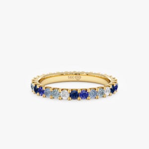 Puede incluir: Un anillo de banda de oro con un patrón repetido de piedras preciosas azules y blancas. El anillo presenta una variedad de tonos azules, incluyendo azul claro, azul oscuro y piedras blancas. El anillo está marcado con "14K".