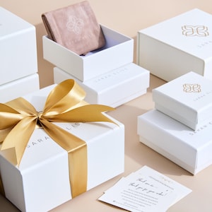 May include: A collection of white jewellery gift boxes with gold logos and a gold ribbon. One box is open, revealing a dusty pink velvet pouch. A small white card with black text is placed in the foreground, featuring a thank you message.