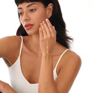 May include: A delicate gold hand chain featuring four small, round, white gemstones. The chain extends from the ring finger to the wrist, creating a minimalist and elegant look. The jewelry is worn on a person's hand.