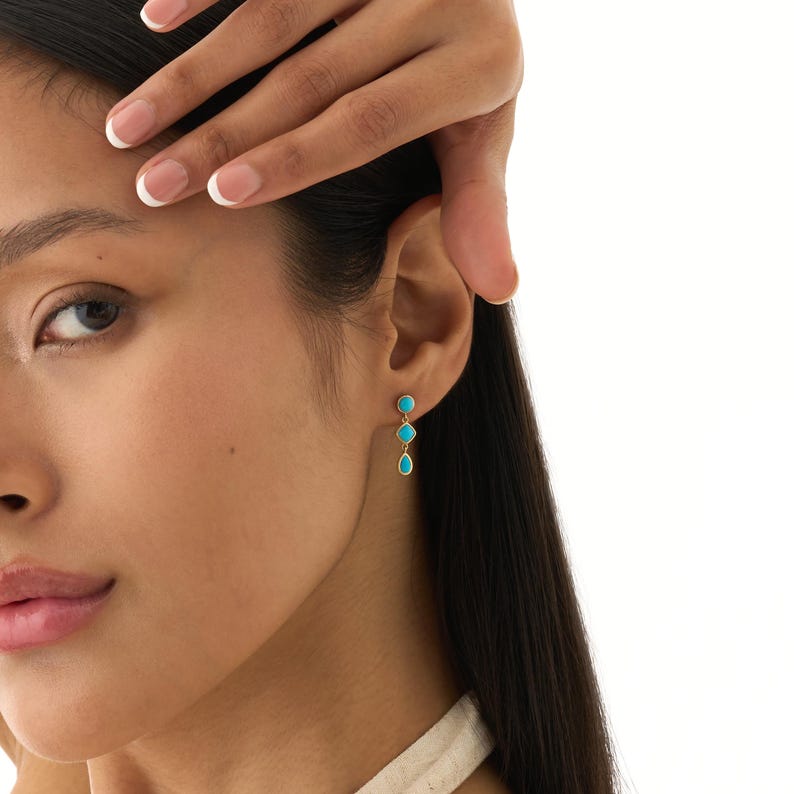 May include: Gold-toned earrings with three turquoise-colored stones. The earrings are dangling and feature a teardrop shape at the bottom. The earrings are worn on a person's ear.