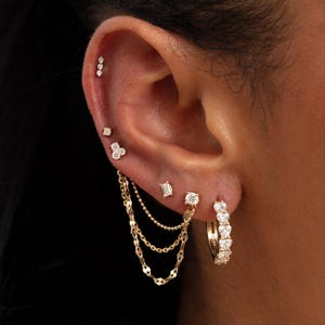 Könnte beinhalten: Nahaufnahme eines Ohrs, das mit mehreren goldenen Ohrringen geschmückt ist. Das Ohr zeigt eine Reihe kleiner, funkelnder Ohrstecker, einen Kettenohrring und einen Creolenohrring mit herzförmigen Edelsteinen. Der Schmuck ist vor einem dunklen Hintergrund.