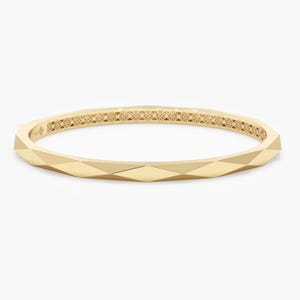 Puede incluir: Un brazalete dorado con un diseño geométrico. El brazalete tiene un exterior facetado y un patrón interior decorativo. El diseño es moderno y elegante, adecuado para el uso diario o ocasiones especiales.