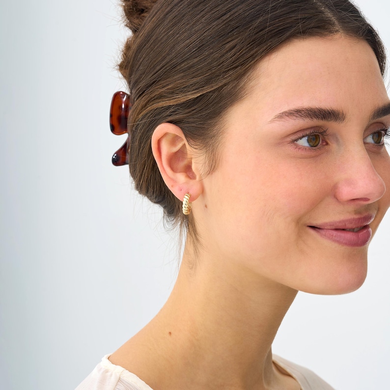 May include: Gold-colored hoop earrings with a twisted rope design. The earrings are small and worn on the ear. The image shows a person wearing the earrings, with a brown hair clip in their hair.