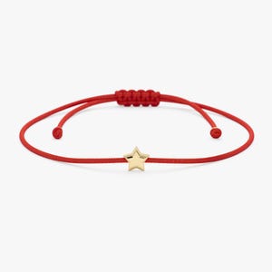 Puede incluir: Pulsera roja ajustable con un dije de estrella dorada. La pulsera tiene un cordón rojo tejido y un cierre de nudo corredizo. El dije es una estrella de cinco puntas.