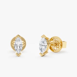 May include: A pair of gold stud earrings featuring a marquise-cut diamond. The diamonds are set in a bezel setting with a decorative backing. The earrings are displayed against a white background.
