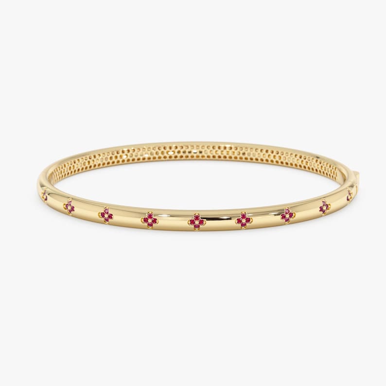 May include: A polished gold bangle bracelet featuring a series of evenly spaced, small four-petalled flower motifs. Each flower is set with tiny, vibrant red stones, and the inner surface of the bangle displays a fine, repeating honeycomb pattern.